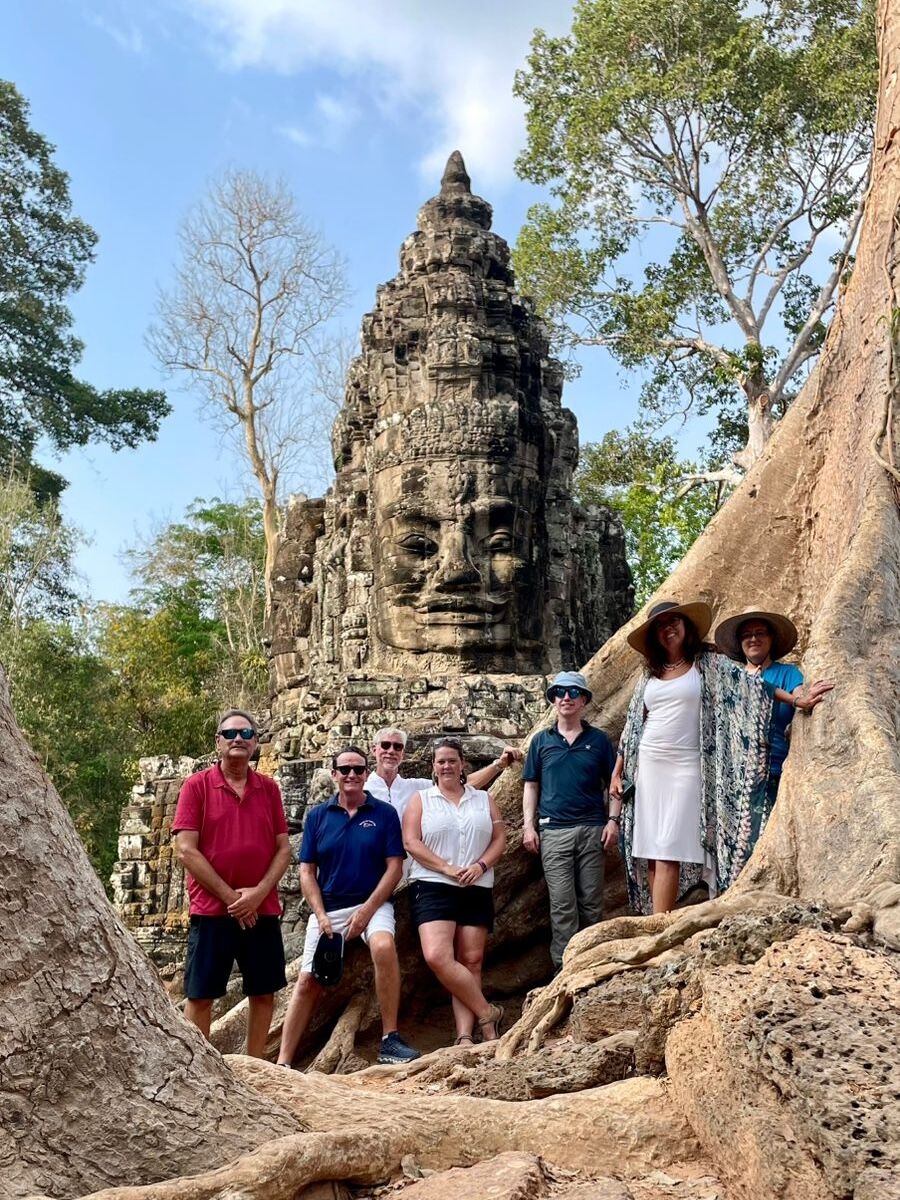 Group of tourists posing in front of a stone face tower at Angkor Thom, Cambodia, surrounded by tree roots and jungle scenery