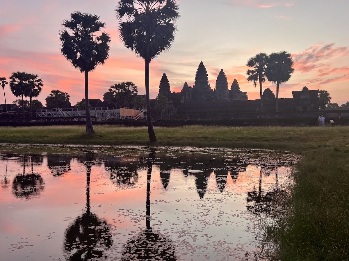 Angkor Wat temple at sunrise with vibrant pink and orange sky reflecting in a lotus pond, framed by palm trees in Cambodia