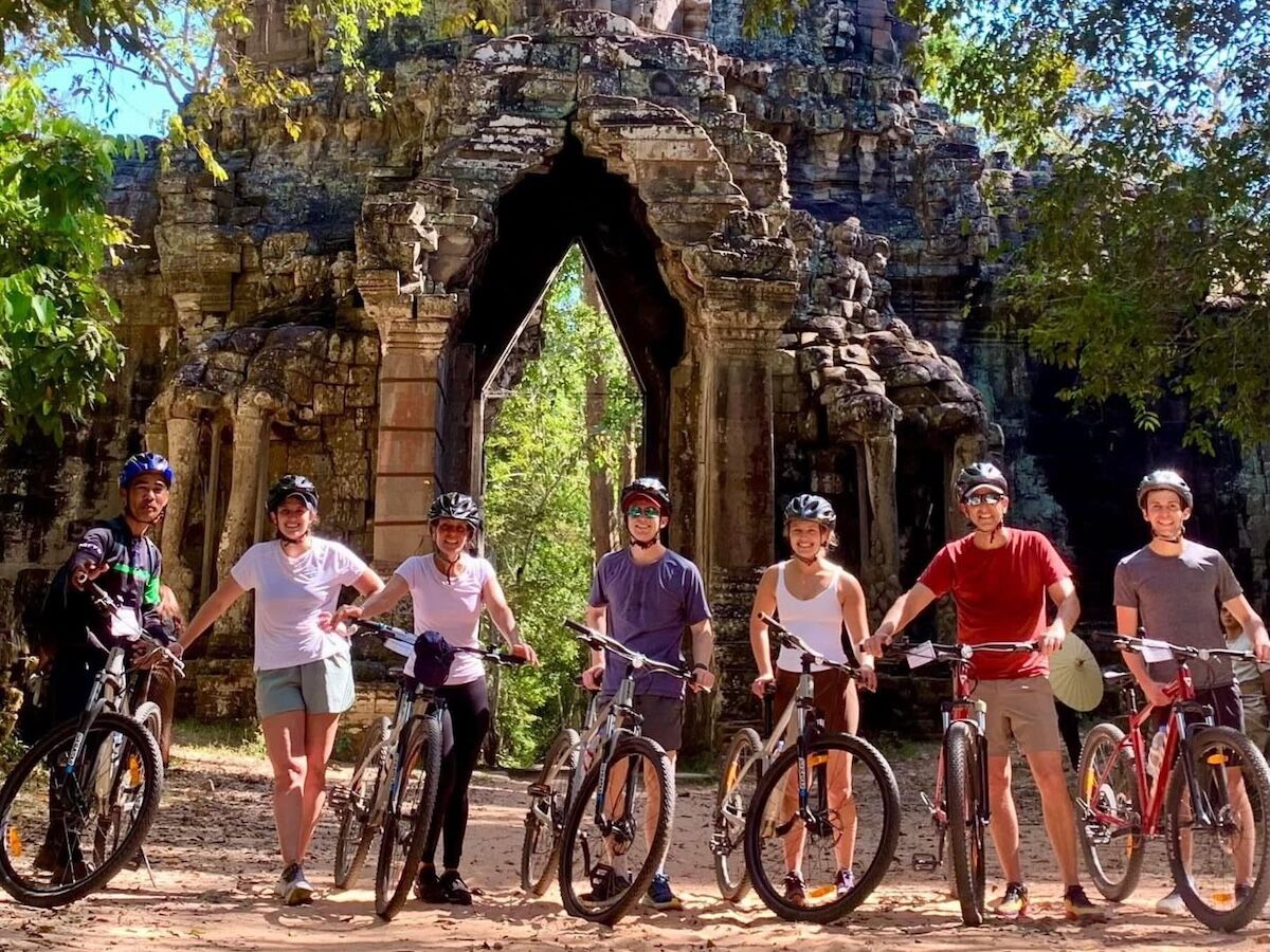 Group of cyclists posing with their bikes in front of an ancient stone gate at Angkor, Cambodia, during a guided temple bike tour