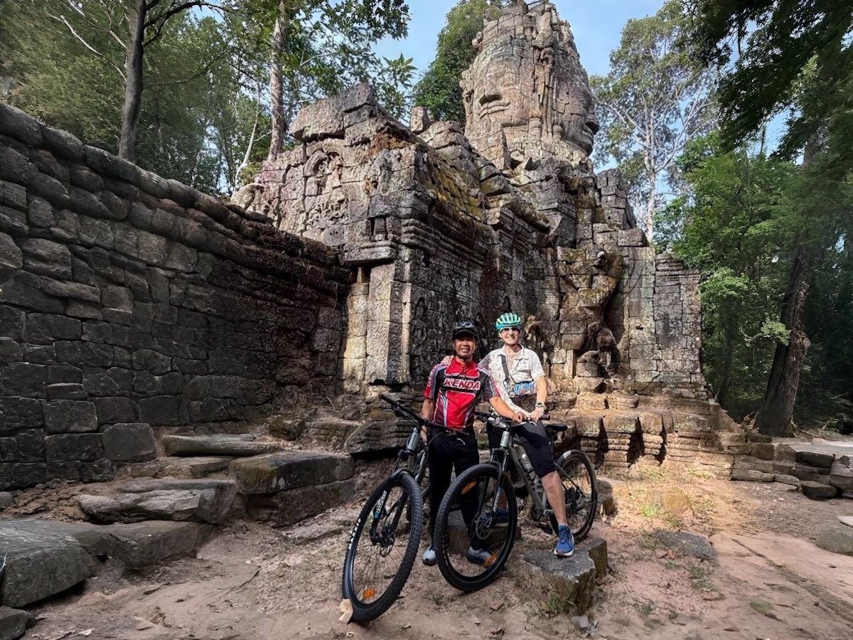 Two cyclists posing with mountain bikes in front of a Bayon-style stone face gate in Angkor Thom, surrounded by ancient ruins and forest
