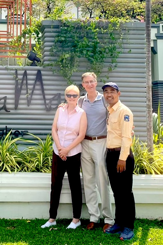 Happy Guests with Local Tour Guide at Cambodia Backroads Tour Start Point Smiling couple posing with their Cambodian tour guide in front of a modern sign and garden area, ready to begin their Cambodia Backroads adventure