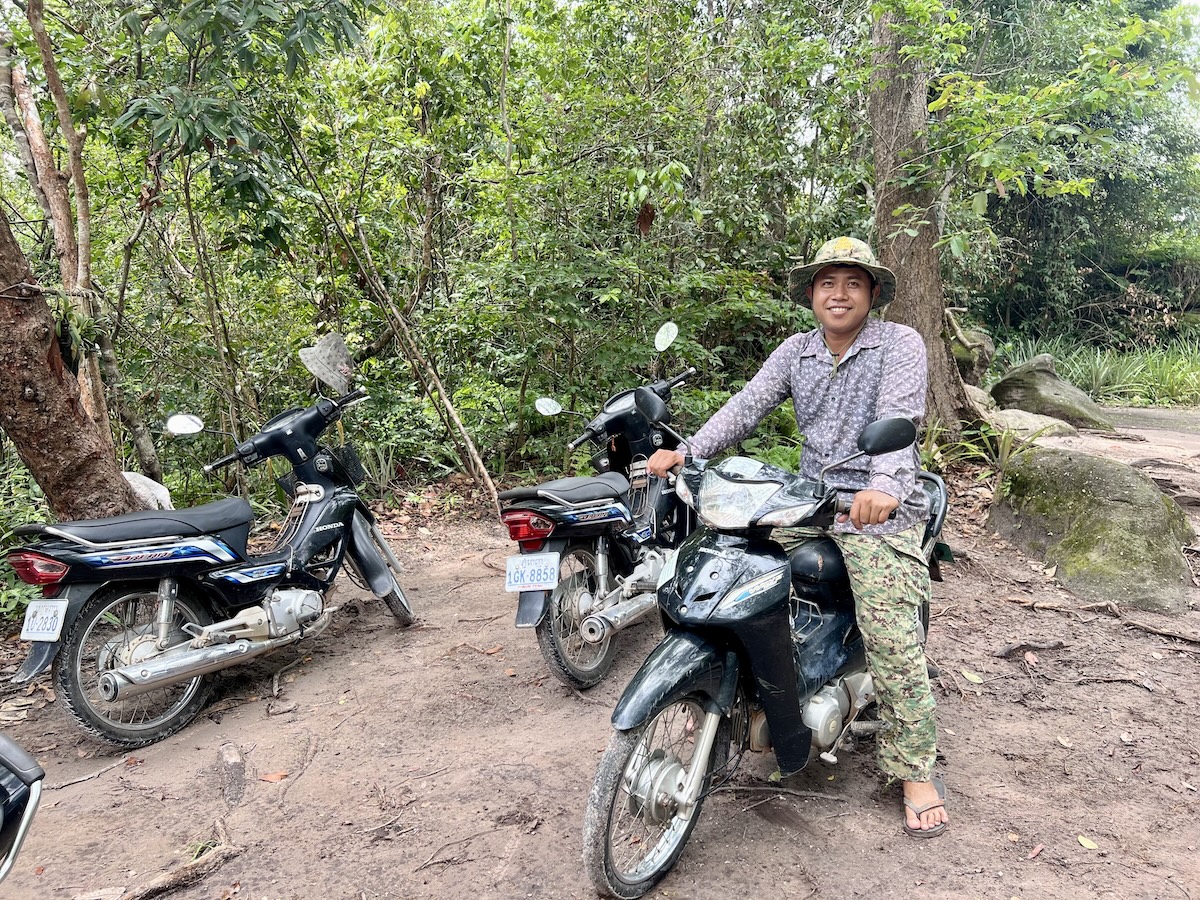 Smiling Cambodian guide sitting on a motorbike in a forested area of Phnom Kulen, surrounded by other parked bikes and tropical greenery