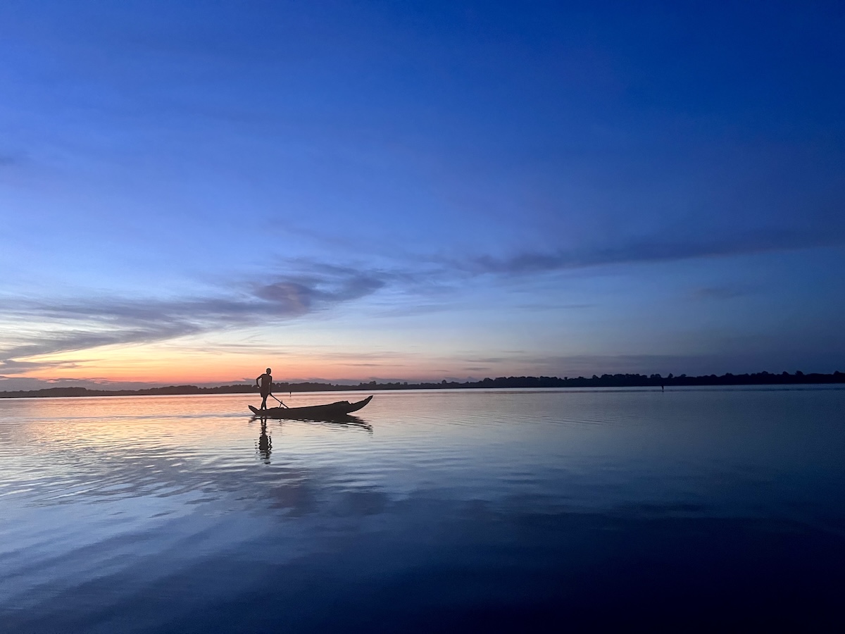 Silhouette of a fisherman gliding a wooden boat across the calm waters of West Baray near Angkor, Cambodia, as the sky glows with sunset colors.