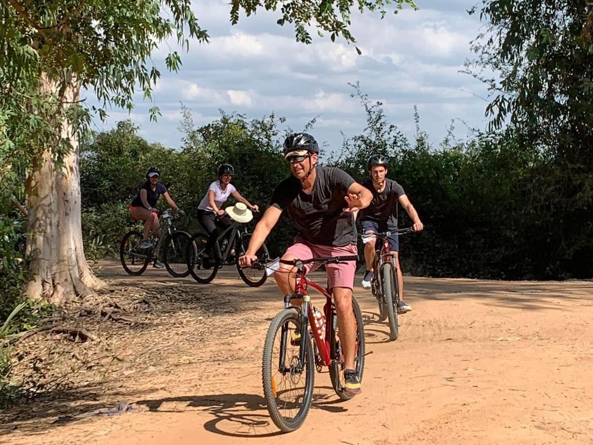 Group of travelers cycling on a sandy backroad in rural Cambodia, enjoying a guided bike tour through scenic countryside with trees and open sky.