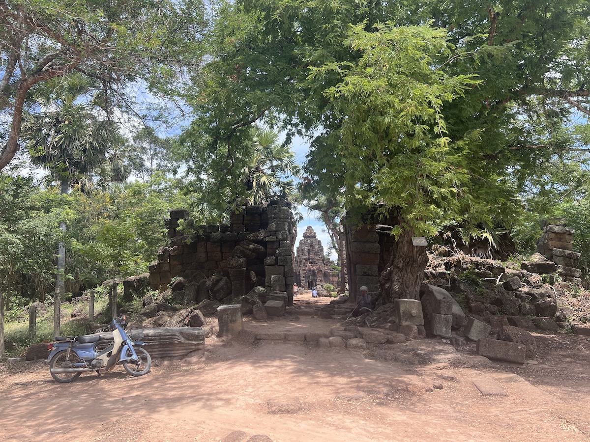 Laterite stone gateway ruins and shaded dirt path leading toward the brick temple tower of Phnom Chisor in Takeo Province, Cambodia, with trees and a parked motorbike in the foreground.