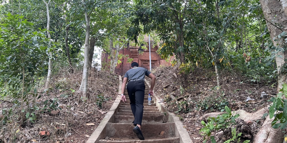 A man ascending steep stone steps through forest toward the red brick pre-Angkorian temple of Phnom Da in Takeo Province, Cambodia.