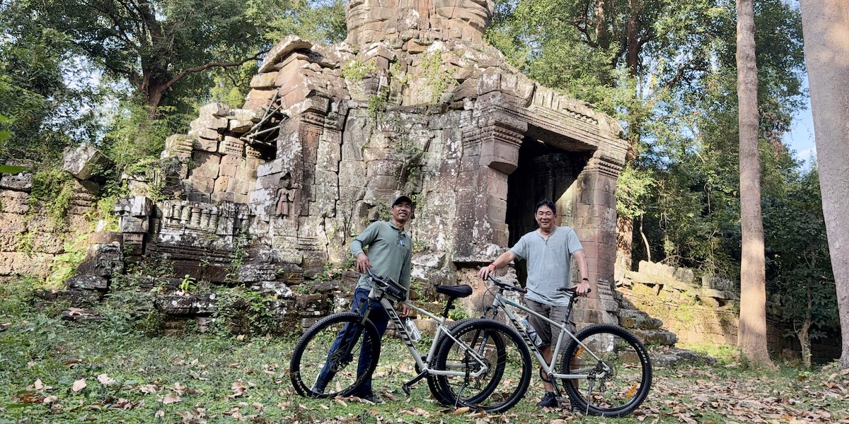 Two men standing with mountain bikes in front of the partially ruined sandstone sanctuary of Ta Som temple in Angkor Archaeological Park, surrounded by trees and fallen leaves.