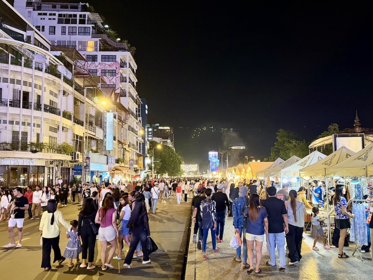 Crowds walking between brightly lit market stalls and riverside buildings at Chaktomuk Walk Street in Phnom Penh, Cambodia.
